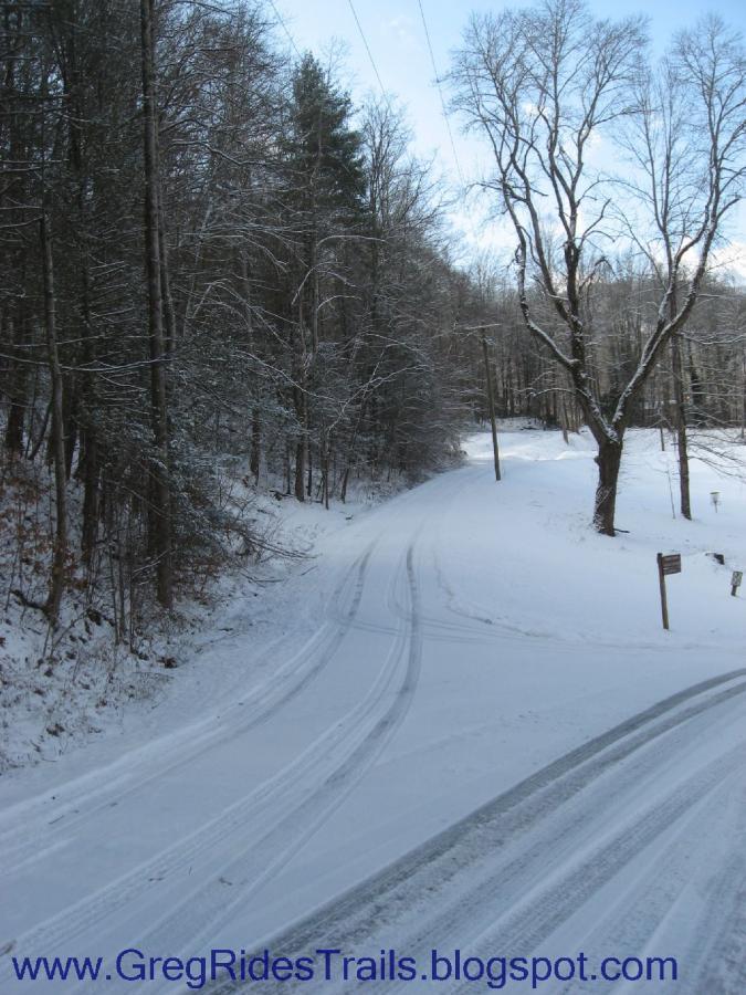 A snow-covered road winding through a winter landscape, flanked by trees with bare branches and evergreen foliage. The scene is peaceful, with tracks visible in the fresh snow leading off into the distance. The sky is partly cloudy, hinting at clear weather. A signpost can be seen on the right side of the road. Fontana Village mountain bike trail.
