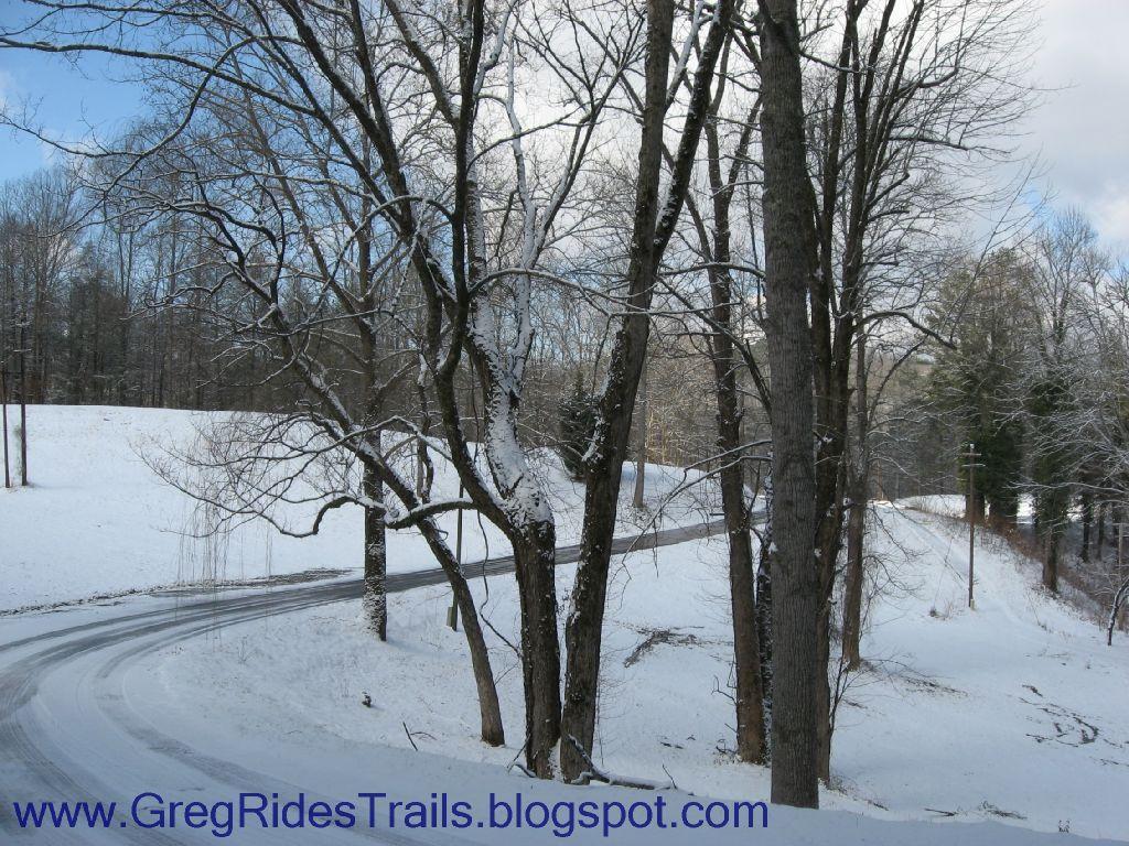 A winter scene featuring bare trees covered with a light dusting of snow, set against a backdrop of a snowy landscape and a winding road. The sky is partly cloudy, with patches of blue visible. Fontana Village mountain bike trail.