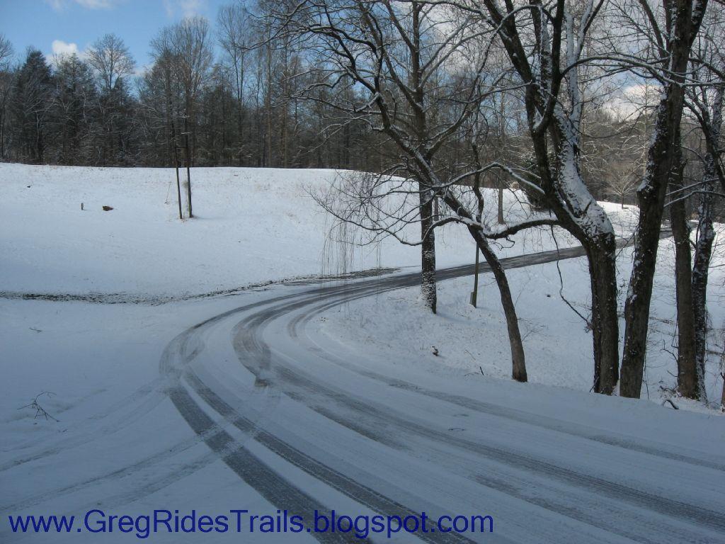 A snowy landscape with a winding road curving through it. Bare trees line the road, and a serene, snowy field is visible in the background under a partly cloudy sky. The scene captures the beauty of a winter day. Fontana Village mountain bike trail.