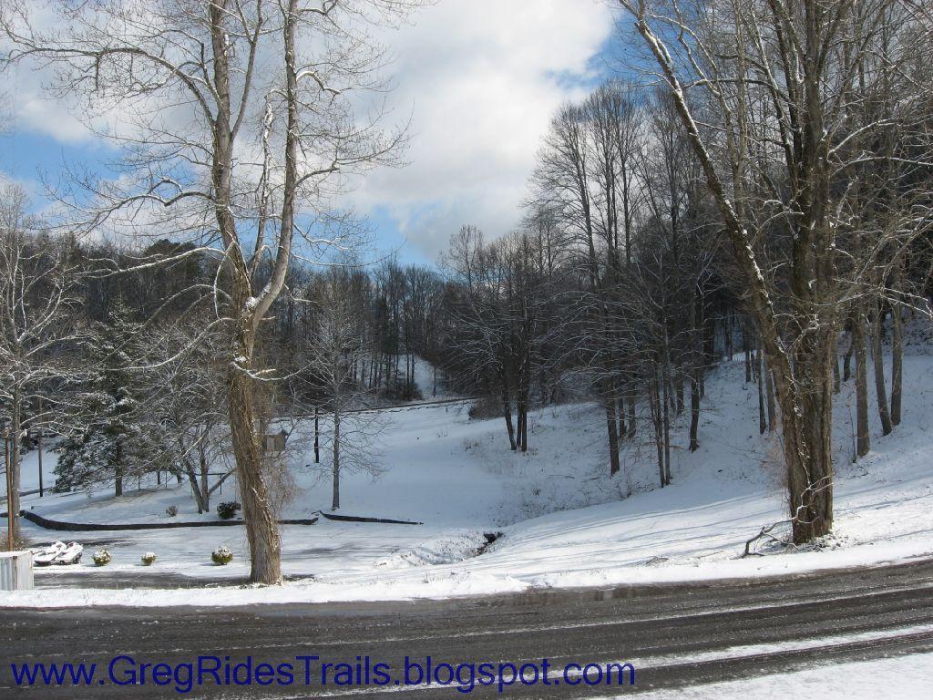 A winter landscape featuring a snow-covered field bordered by tall trees and a winding road in the foreground. The sky is partly cloudy, with patches of blue peeking through. Snow blankets the ground, creating a serene, tranquil atmosphere. Fontana Village mountain bike trail.
