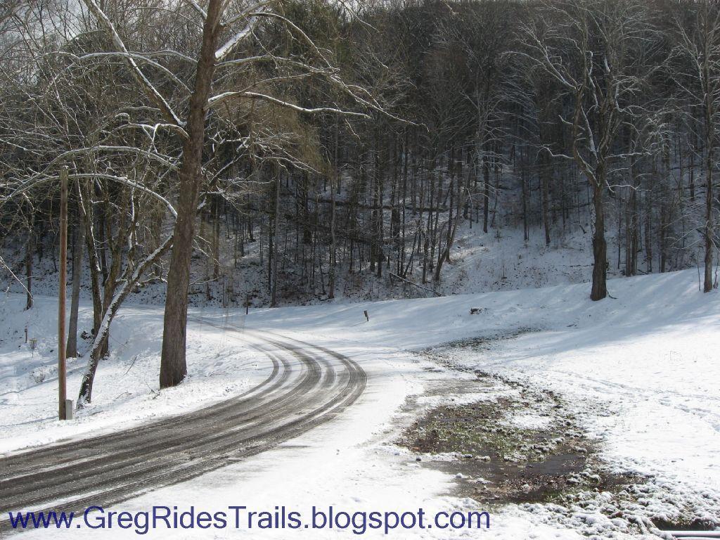 A snowy landscape featuring a winding road cutting through a wintry scene. Tall, bare trees line the sides of the road, with patches of snow covering the ground. The scene captures the serene beauty of winter in a wooded area. Fontana Village mountain bike trail.