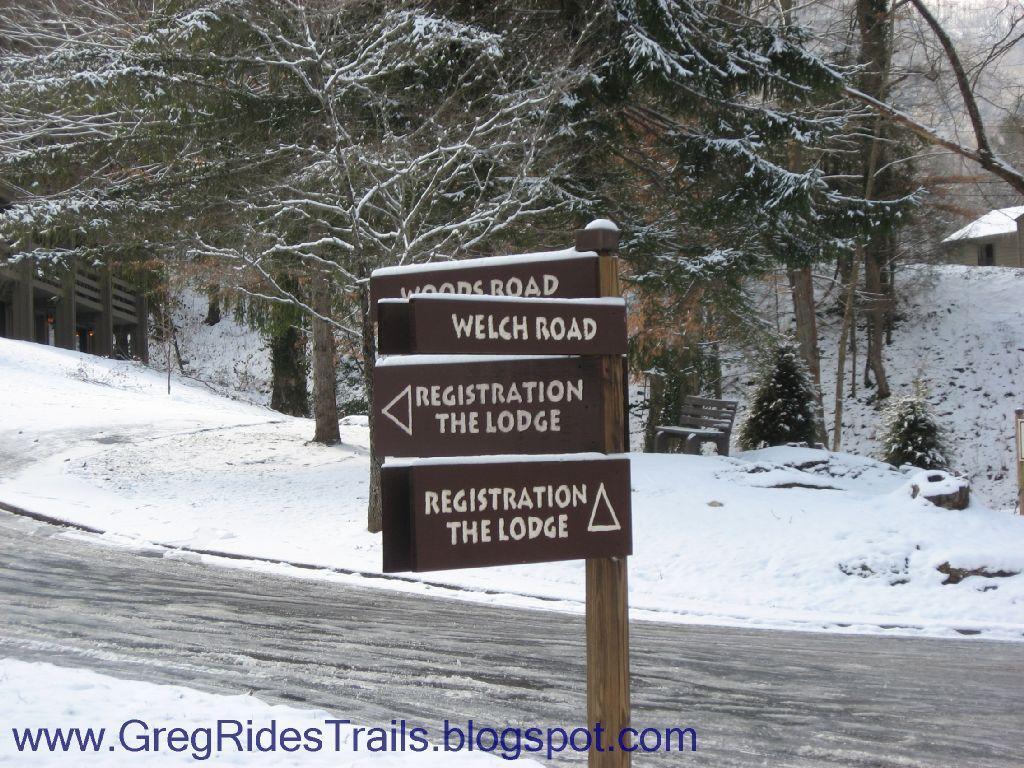 Snow-covered road signs at a winter lodge, indicating directions to Woodes Road, Welch Road, and Registration for the Lodge. Pine trees are visible in the background, with a light layer of snow covering the ground and surrounding areas. Fontana Village mountain bike trail.