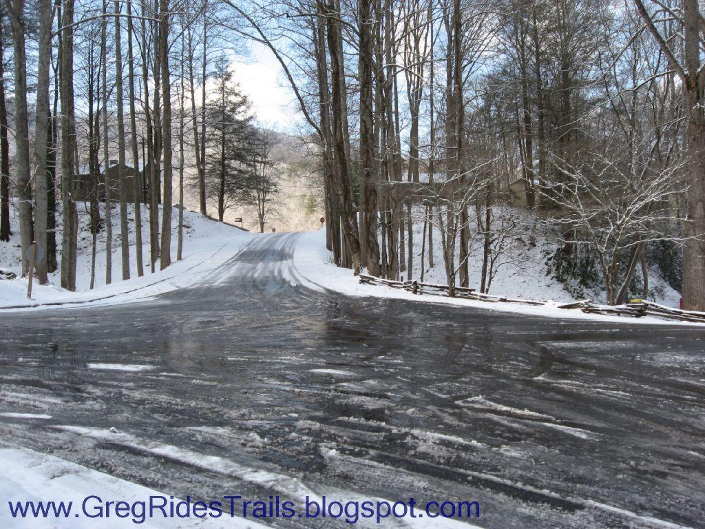 A snowy road winding through a wooded area, with trees on either side and a slight incline in the background. The road appears icy and shiny, reflecting the sky above, which is partly cloudy. Snow covers the ground and branches, creating a serene winter landscape. Fontana Village mountain bike trail.