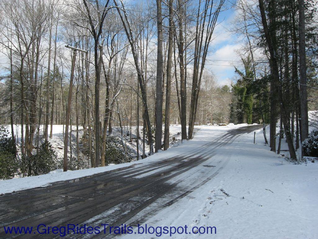 A snow-covered path surrounded by bare trees, with bright blue skies peeking through scattered clouds. The road stretches ahead, winding gently through a serene winter landscape. Fontana Village mountain bike trail.