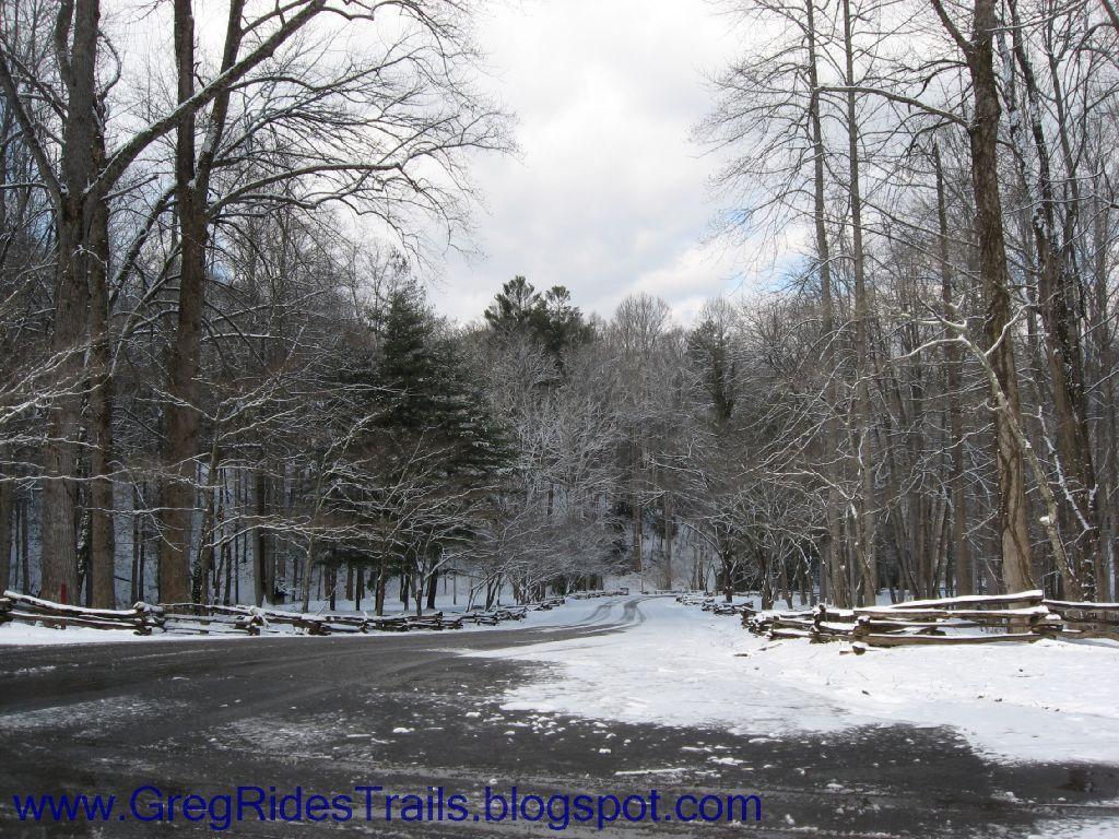 A scenic winter landscape featuring a snow-covered road winding through a dense forest of bare and evergreen trees. The ground is blanketed in fresh snow, and a rustic wooden fence lines the road, enhancing the tranquil winter atmosphere under a cloudy sky. Fontana Village mountain bike trail.
