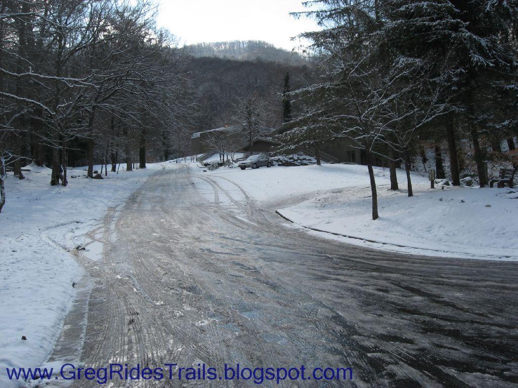 A snow-covered road winding through a scenic winter landscape, flanked by bare trees and a distant view of mountains. A few buildings are visible along the roadside, with patches of ice reflecting the cold sunlight. The scene evokes a tranquil, wintry atmosphere. Fontana Village mountain bike trail.