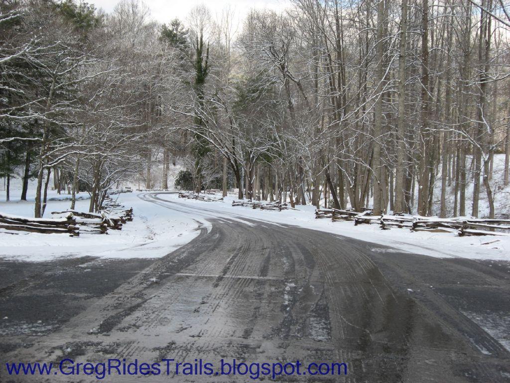 A winding, snow-covered road surrounded by bare trees and a wooden fence, capturing a tranquil winter scene. The landscape features a gentle curve in the road, with fresh snow blanketing the ground and branches, creating a serene atmosphere. Fontana Village mountain bike trail.