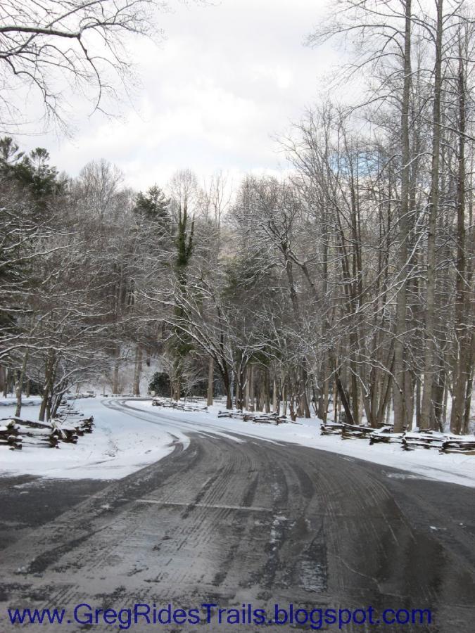 A snowy road winding through a forest, lined with bare trees and a wooden fence, under a cloudy sky. The scene captures the serenity of winter in a wooded area. Fontana Village mountain bike trail.