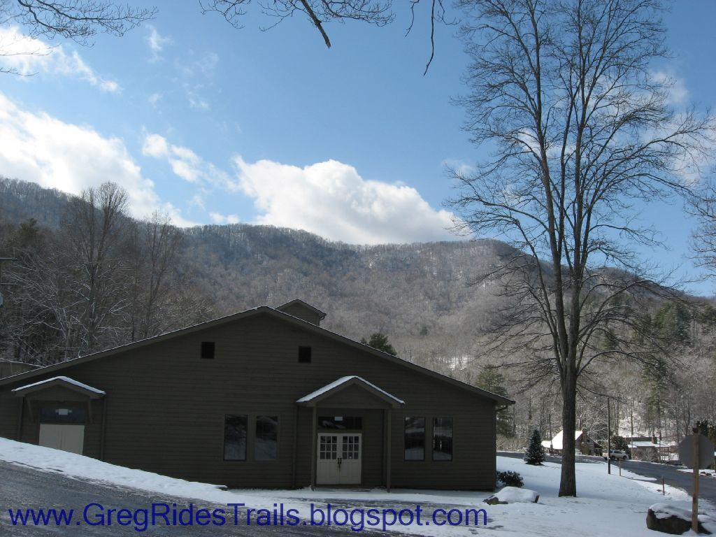 A brown building partially covered in snow, set against a backdrop of mountains under a cloudy blue sky. Leafless trees are visible in the foreground, and the ground is blanketed in fresh snow. The scene conveys a serene winter landscape. Fontana Village mountain bike trail.