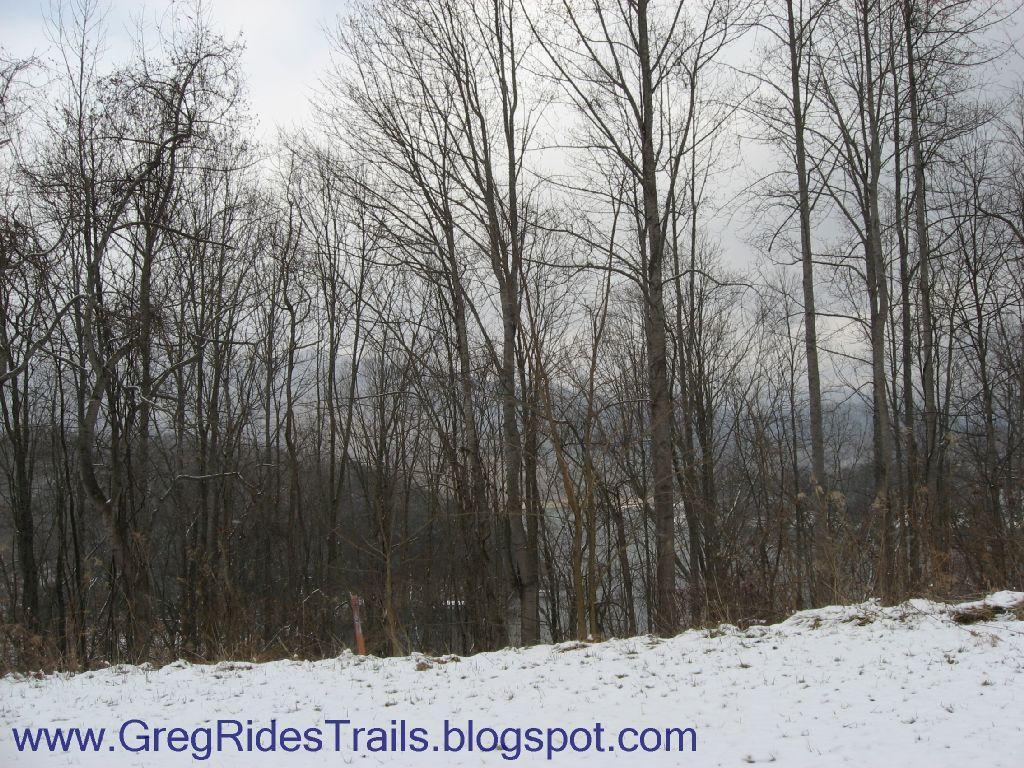 A winter landscape featuring bare trees with a light dusting of snow on the ground, set against a cloudy sky. In the background, a glimpse of water can be seen through the trees. The scene evokes a tranquil, wintry atmosphere. Fontana Village mountain bike trail.