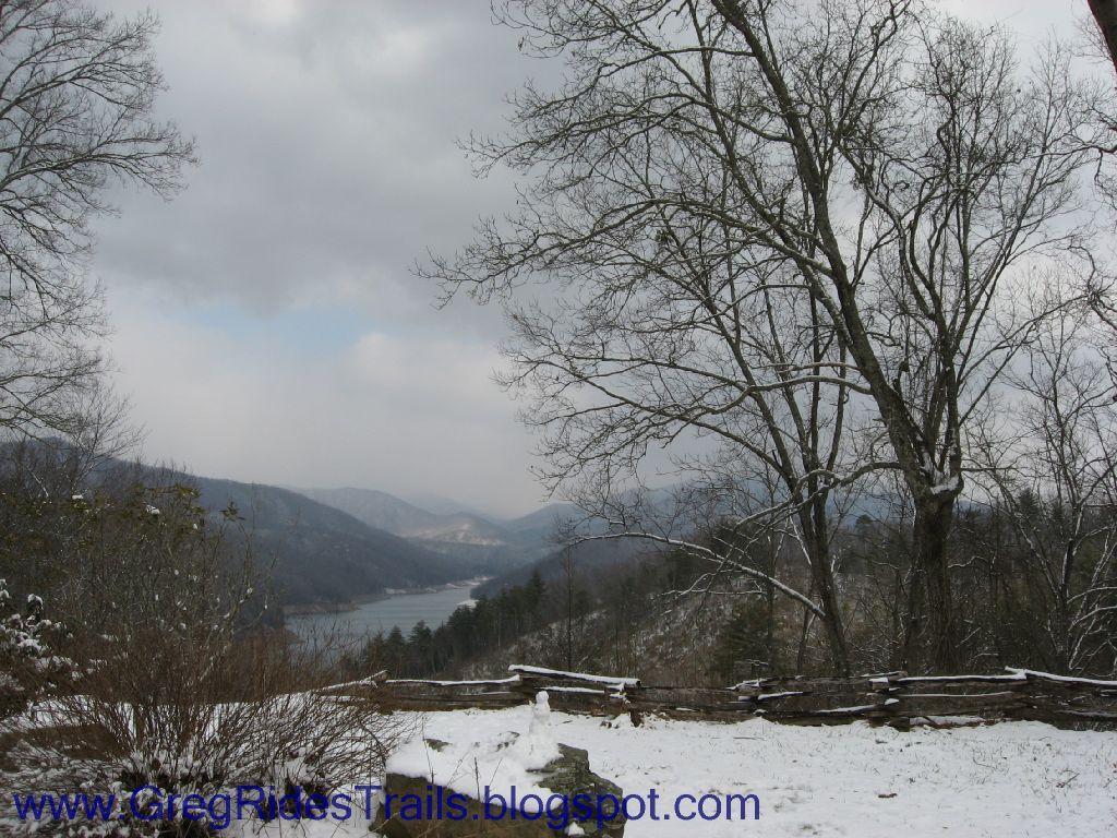 A scenic view of a snowy landscape featuring a lake surrounded by mountains, with a foreground of leafless trees and a rustic wooden fence. The sky is overcast, creating a somber yet tranquil atmosphere. Fontana Village mountain bike trail.