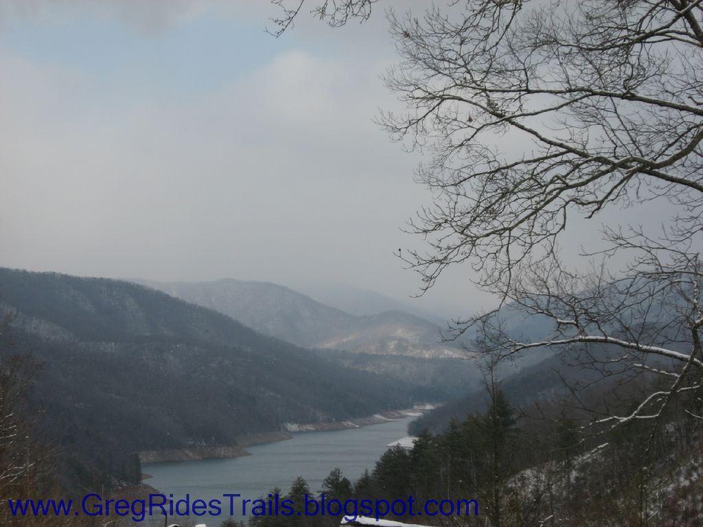 A scenic view of a mountainous landscape with a river winding through it, surrounded by trees and hills. The scene is set under a cloudy sky, with hints of snow on the mountains. Fontana Village mountain bike trail.