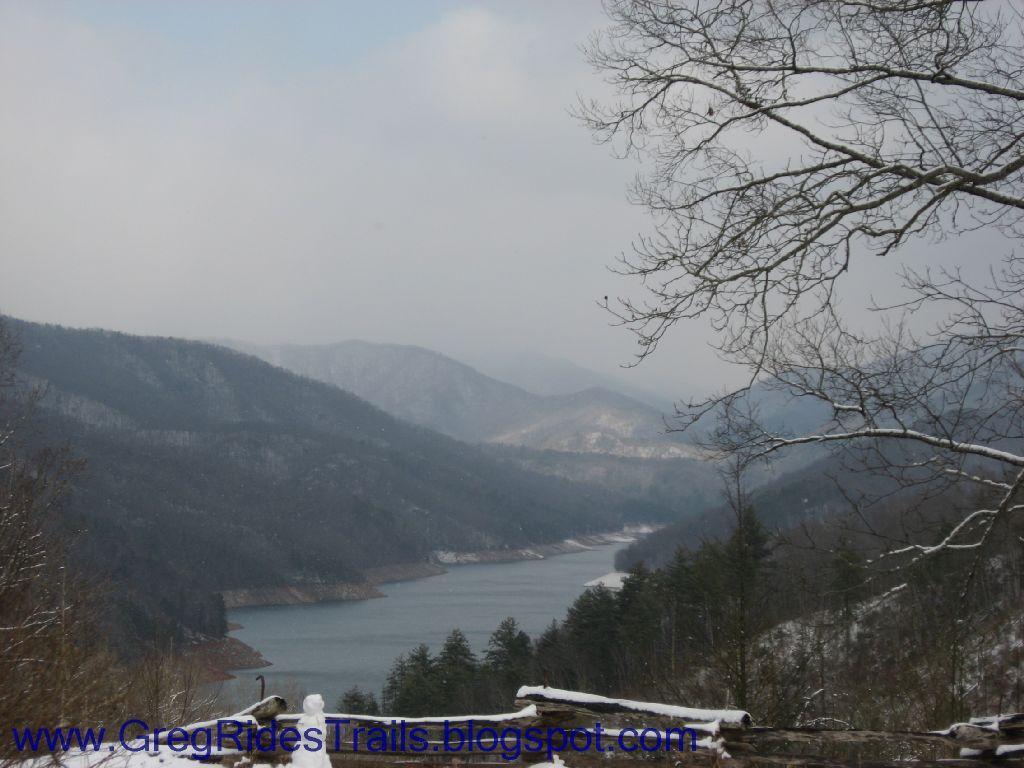 A scenic view of a misty mountain landscape with a calm lake in the foreground, surrounded by tree-lined banks and rolling hills. The scene captures a tranquil winter atmosphere, with hints of snow on the ground and a cloudy sky overhead. Fontana Village mountain bike trail.