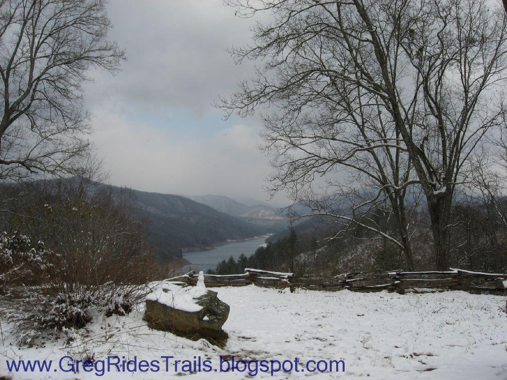 A snowy landscape featuring a view of a lake surrounded by mountains, with bare trees and a stone in the foreground. The sky is overcast, creating a serene winter atmosphere. Fontana Village mountain bike trail.