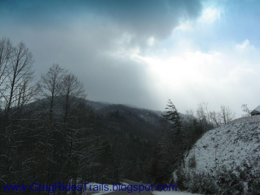 A winter landscape featuring snow-covered mountains and bare trees under a cloudy sky. The scene captures a tranquil and chilly atmosphere, with a hint of sunlight peeking through the clouds in the background. Fontana Village mountain bike trail.