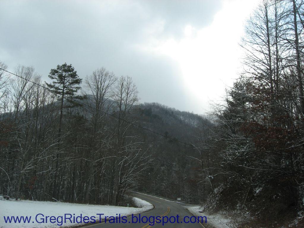A winter landscape showing a curved road surrounded by bare trees covered in a light dusting of snow, leading toward a distant, misty mountain under a cloudy sky. Fontana Village mountain bike trail.