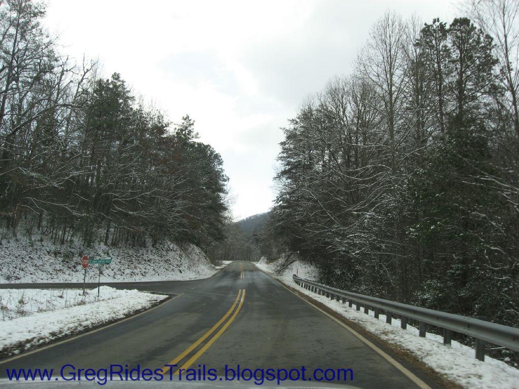A snow-covered road winding through a forest of bare and evergreen trees under a cloudy sky. Fontana Village mountain bike trail.