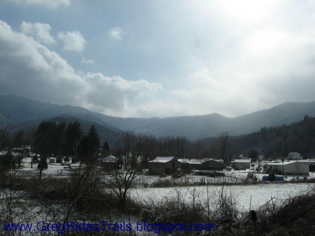A winter landscape featuring a rural area blanketed in snow, with small houses set against a backdrop of trees and mountains under a partly cloudy sky. Fontana Village mountain bike trail.