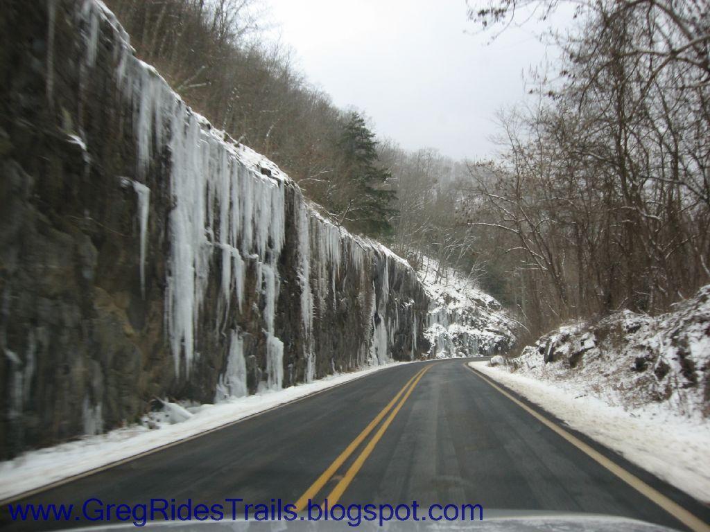 A snowy road winding through a mountainous area, flanked by a rocky cliff adorned with icicles. The landscape is covered in a light dusting of snow, with leafless trees visible in the background under a cloudy sky. Fontana Village mountain bike trail.