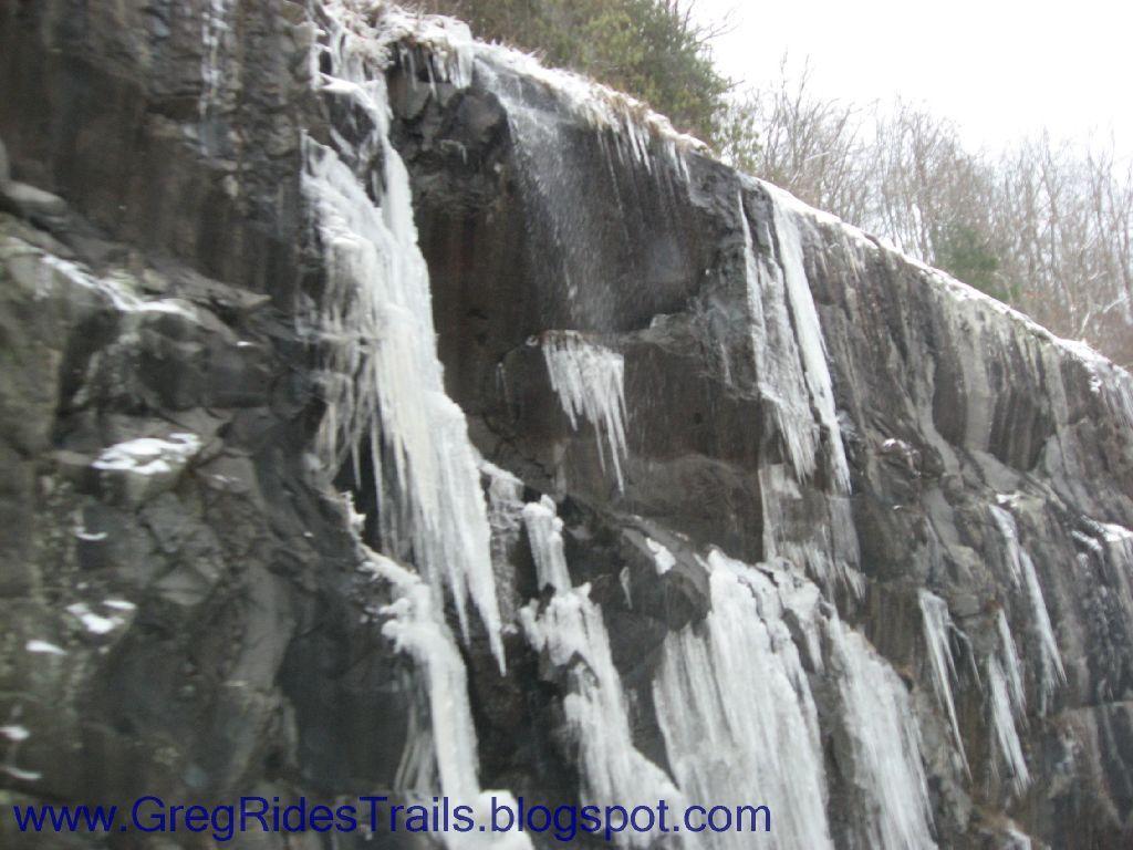 Icicles hanging from a rocky cliff with a light dusting of snow, surrounded by a forested area in winter. Fontana Village mountain bike trail.