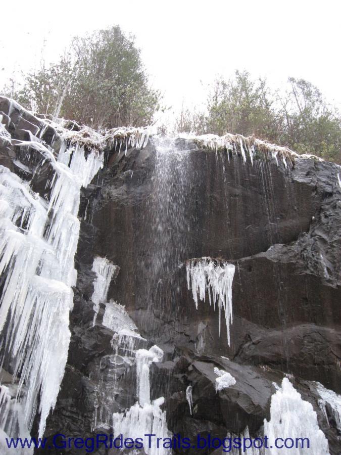 A cascading waterfall is visible, surrounded by dark rock formations, with icicles hanging from the edges. The scene depicts the natural beauty of winter, where water flows over the cliff and drips down, with greenery visible at the top. The overall atmosphere is serene and cold, capturing the essence of a winter landscape. Fontana Village mountain bike trail.