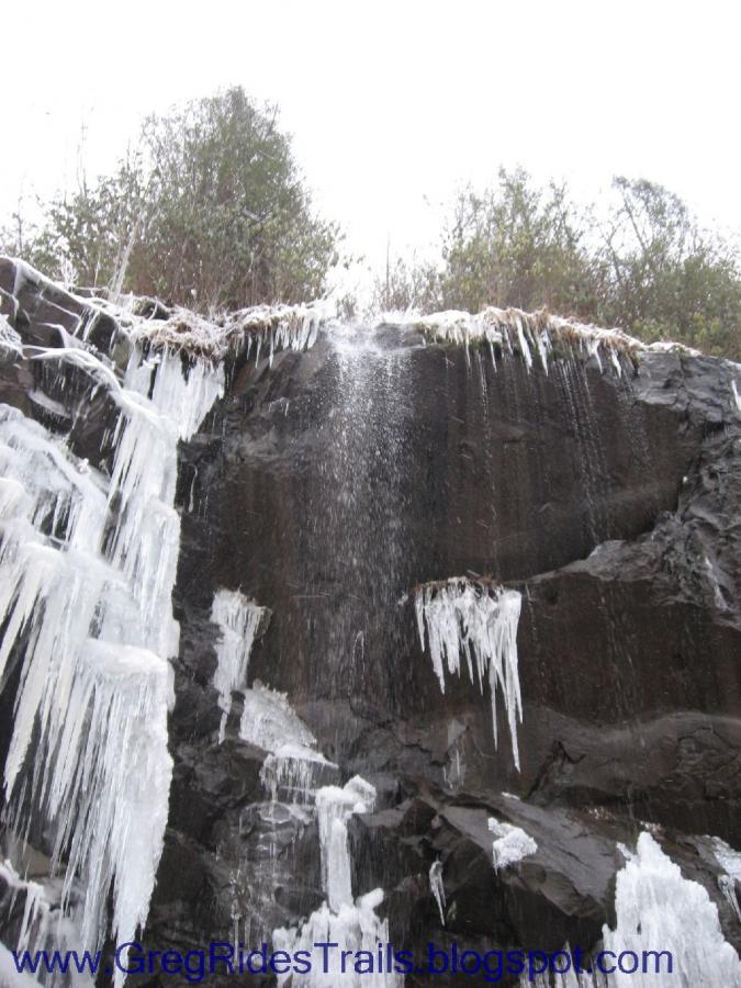 A cascading waterfall surrounded by icy formations on a rocky cliff. The falling water splashes down, while icicles hang from the edges. The background features a cloudy sky and sparse greenery. Fontana Village mountain bike trail.