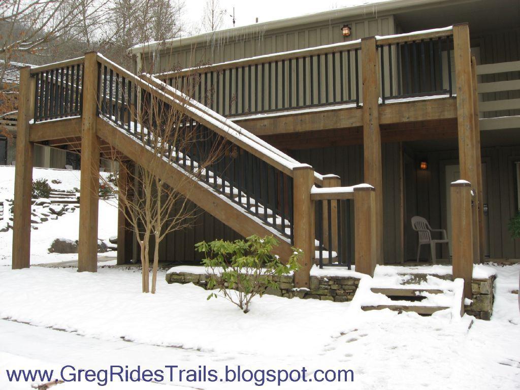Wooden staircase leading to an upper deck, surrounded by a snowy landscape. The scene features a tree and a small shrub at the base of the stairs, along with a stone pathway and a chair near the entrance. The exterior of the building is partially visible in the background. Fontana Village mountain bike trail.