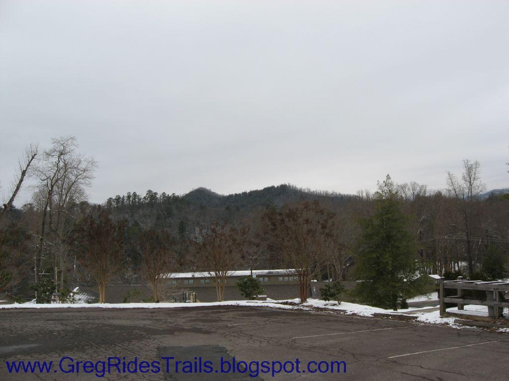A scenic view of a mountainous landscape under a cloudy sky. In the foreground, there is a parking area with patches of snow and bare trees, while the background features rolling hills and a line of trees. The overall atmosphere is tranquil and wintery. Fontana Village mountain bike trail.