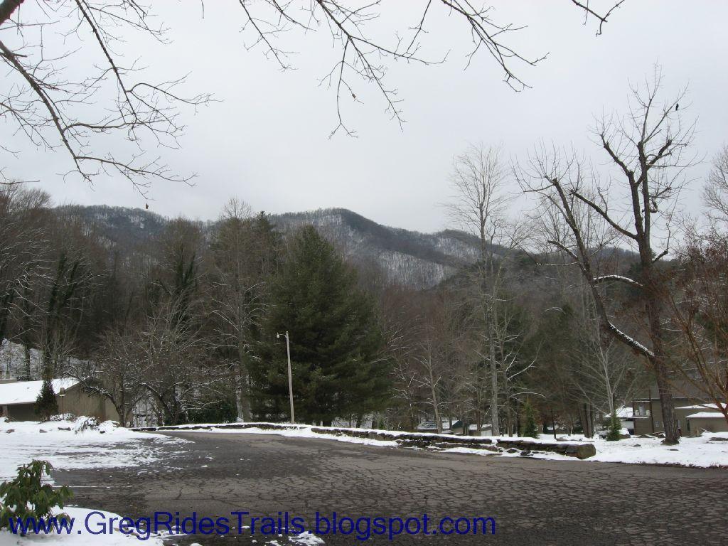 A winter landscape featuring snow-covered ground, trees, and mountains in the background under a cloudy sky. The foreground shows a paved curved road lined with leafless trees and evergreens. Fontana Village mountain bike trail.