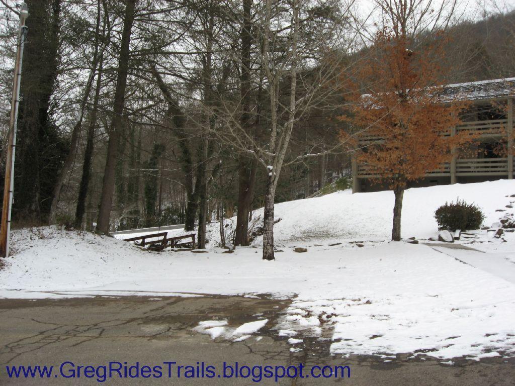 Snow-covered landscape featuring a driveway leading to a wooded area. There are bare trees alongside the path, with a few patches of grass peeking through the snow. A bench is visible in the background, and a building can be seen partially hidden among the trees. The scene conveys a quiet, winter atmosphere. Fontana Village mountain bike trail.
