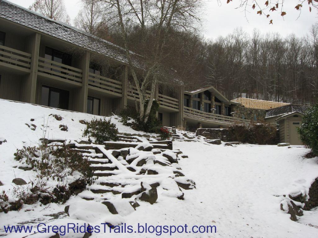 A snow-covered landscape featuring a multi-story lodge with balconies, surrounded by trees. Stone steps lead up from the snowy ground to the entrance of the lodge, set against a backdrop of a winter forest. Fontana Village mountain bike trail.