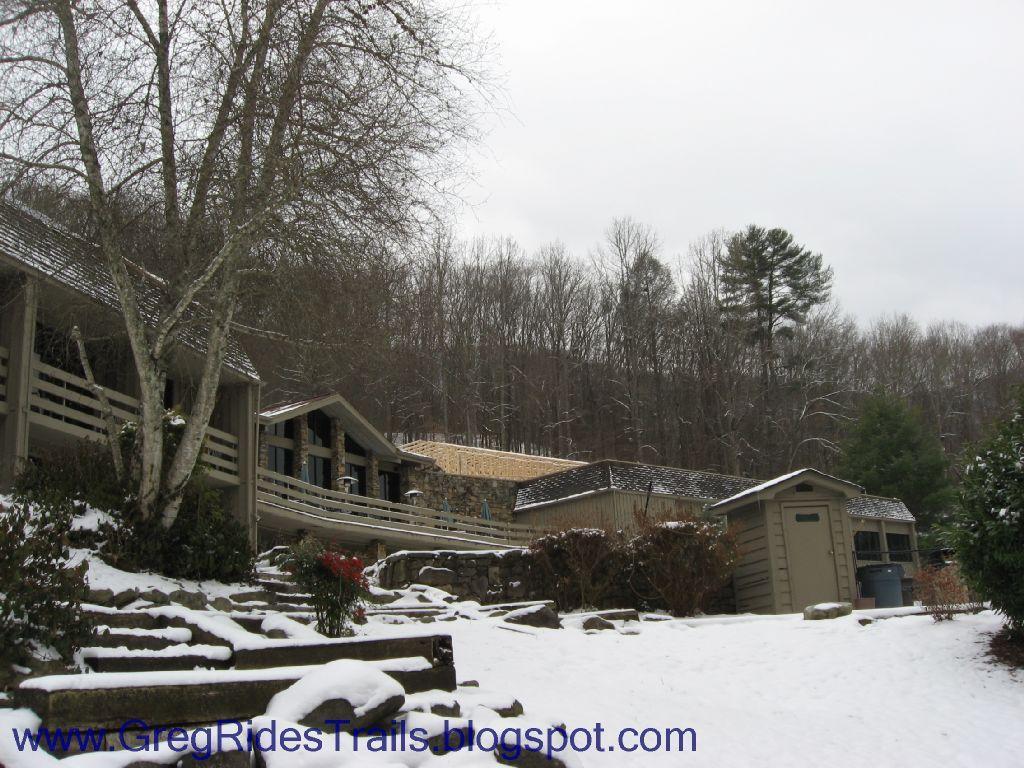 A snow-covered scene featuring several rustic buildings nestled among bare trees. The foreground shows stone steps leading up to the buildings, which have a combination of wooden and stone structures. The landscape is serene, with a gentle winter ambiance, and the sky appears overcast, indicating a chilly day. Fontana Village mountain bike trail.