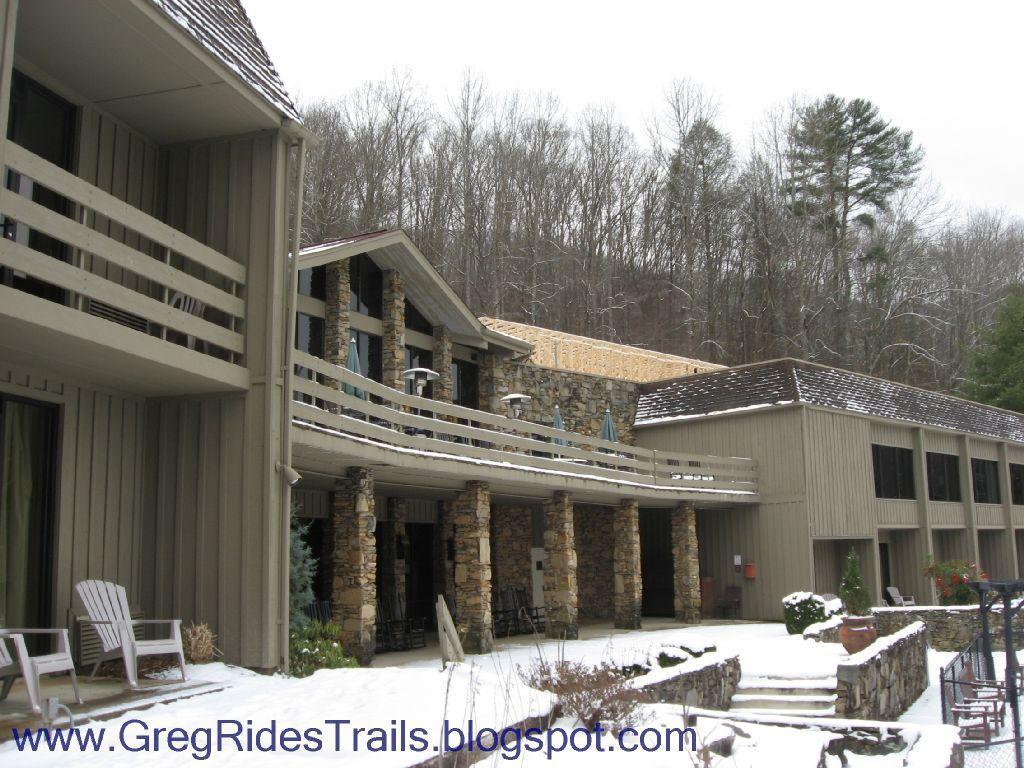 A winter scene of a lodge featuring a combination of wood and stone architecture. Snow covers the ground and the surrounding area, while the building has multiple balconies and large windows. In the foreground, there are wooden chairs and a stone pathway, enhancing the cozy atmosphere amid a backdrop of wooded hills. Fontana Village mountain bike trail.