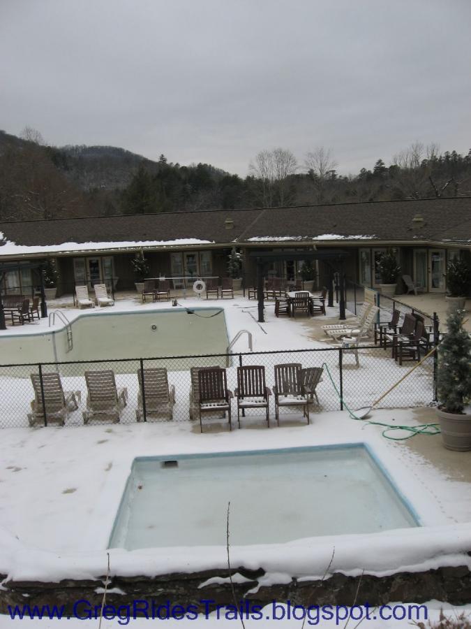 A view of an outdoor pool area surrounded by snow, featuring two swimming pools—one larger and one smaller—alongside several wooden lounge chairs. The area is enclosed by a fence and is set against a backdrop of trees and mountains, with a cloudy sky overhead. Fontana Village mountain bike trail.