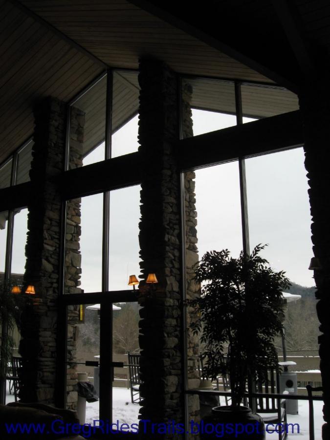 A cozy interior view of a lodge with large windows and stone pillars, showcasing a winter landscape outside. The room features a plant beside the doors and snow visible outside. Soft lighting from sconces adds warmth to the space. Fontana Village mountain bike trail.