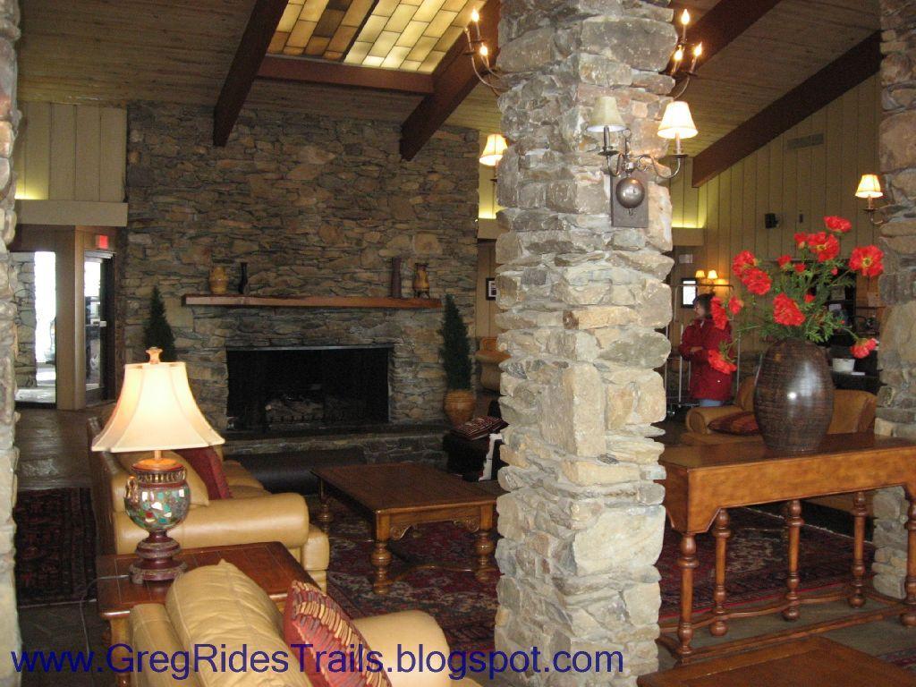 Cozy lodge interior featuring a stone fireplace, warm lighting, and comfortable seating. The space includes wooden beams, stone columns, and decorative floral arrangements, creating a welcoming atmosphere. Fontana Village mountain bike trail.