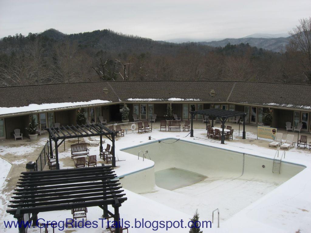 An empty swimming pool surrounded by a snow-covered courtyard and mountains in the background. The scene includes several wooden lounge chairs and a trellis structure, set against a cloudy sky. Fontana Village mountain bike trail.