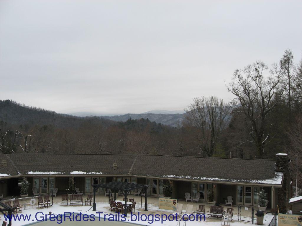 A scenic view of a lodge surrounded by mountains, with a foreground featuring patio furniture and decorative trees. The sky is overcast, and the landscape shows hints of snow on the ground, creating a tranquil, wintry atmosphere. Fontana Village mountain bike trail.