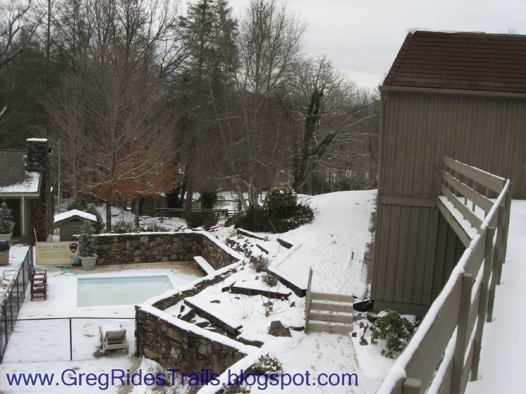 A winter scene showing a snow-covered backyard with a swimming pool partially visible. Surrounding trees are bare, indicating the season, while a wooden structure is situated on the right side of the image. A sign with pool rules is prominently displayed near the pool area. Snow blankets the ground and pathways, creating a serene, quiet atmosphere. Fontana Village mountain bike trail.