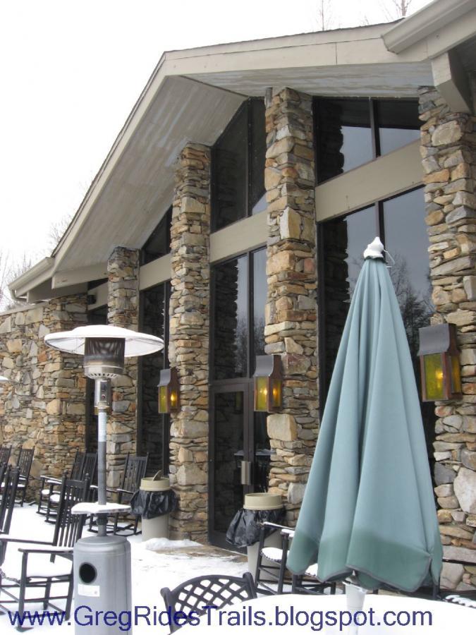 Exterior view of a stone-clad building featuring large glass windows, outdoor seating with black chairs, and a teal umbrella. A patio heater is positioned nearby, and snow can be seen on the ground, suggesting a winter setting. Fontana Village mountain bike trail.