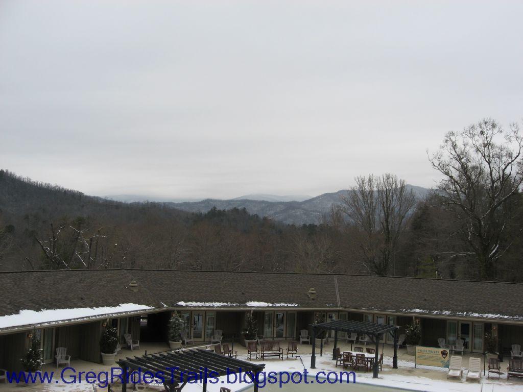 A scenic view of a mountain landscape under a cloudy sky, featuring a low building in the foreground with a terrace area. Snow can be seen on the roof and surrounding ground, while the distant mountains are partially shrouded in mist. Fontana Village mountain bike trail.
