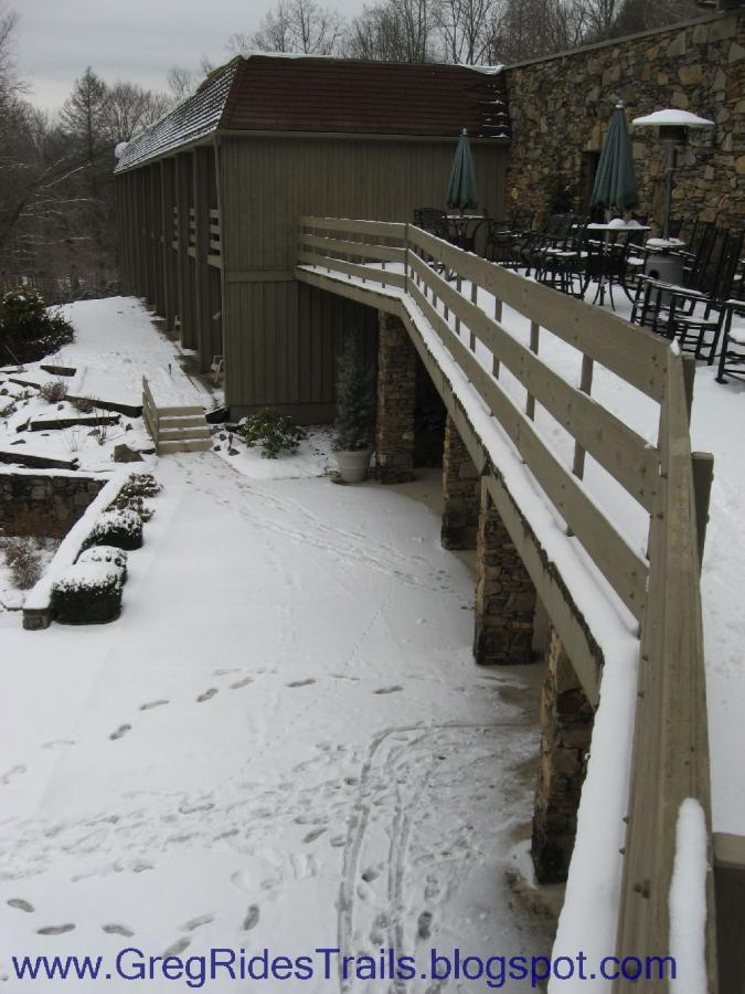 A snow-covered pathway leading to a building, featuring a wooden bridge. The setting includes stone walls and outdoor seating with umbrellas. Footprints are visible in the snow, while surrounding trees are lightly dusted with snowflakes. Fontana Village mountain bike trail.