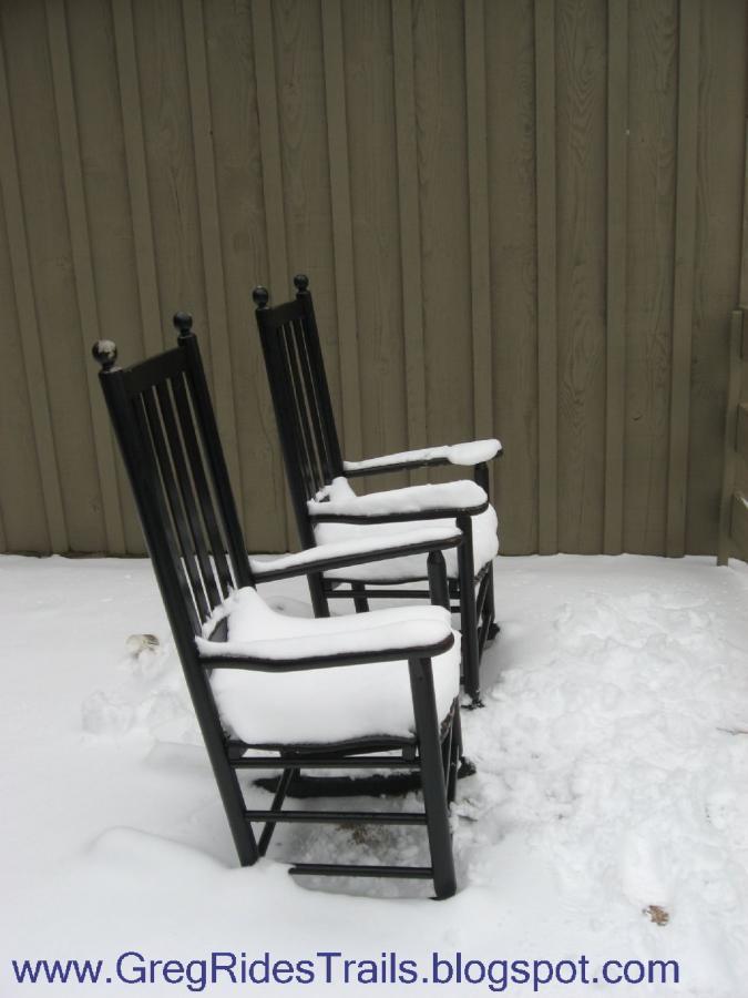 Two black rocking chairs covered with snow, positioned side by side on a snowy ground. A textured wooden wall serves as the background, hinting at a cozy outdoor space. Fontana Village mountain bike trail.