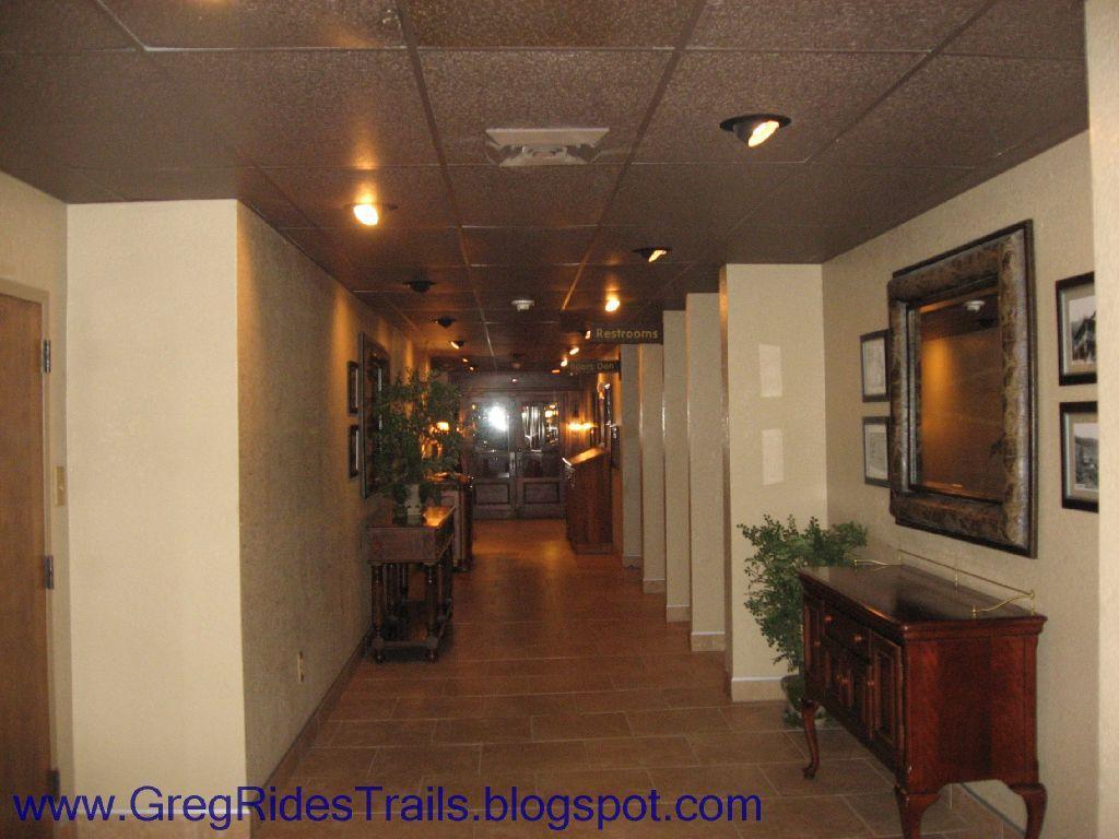 A well-lit hallway featuring tile flooring and textured walls, with doors on the left side leading to restrooms. There are framed pictures on the right wall, and a decorative wooden cabinet with a plant on top is located near the end of the corridor. The hallway appears inviting and is designed for indoor use. Fontana Village mountain bike trail.
