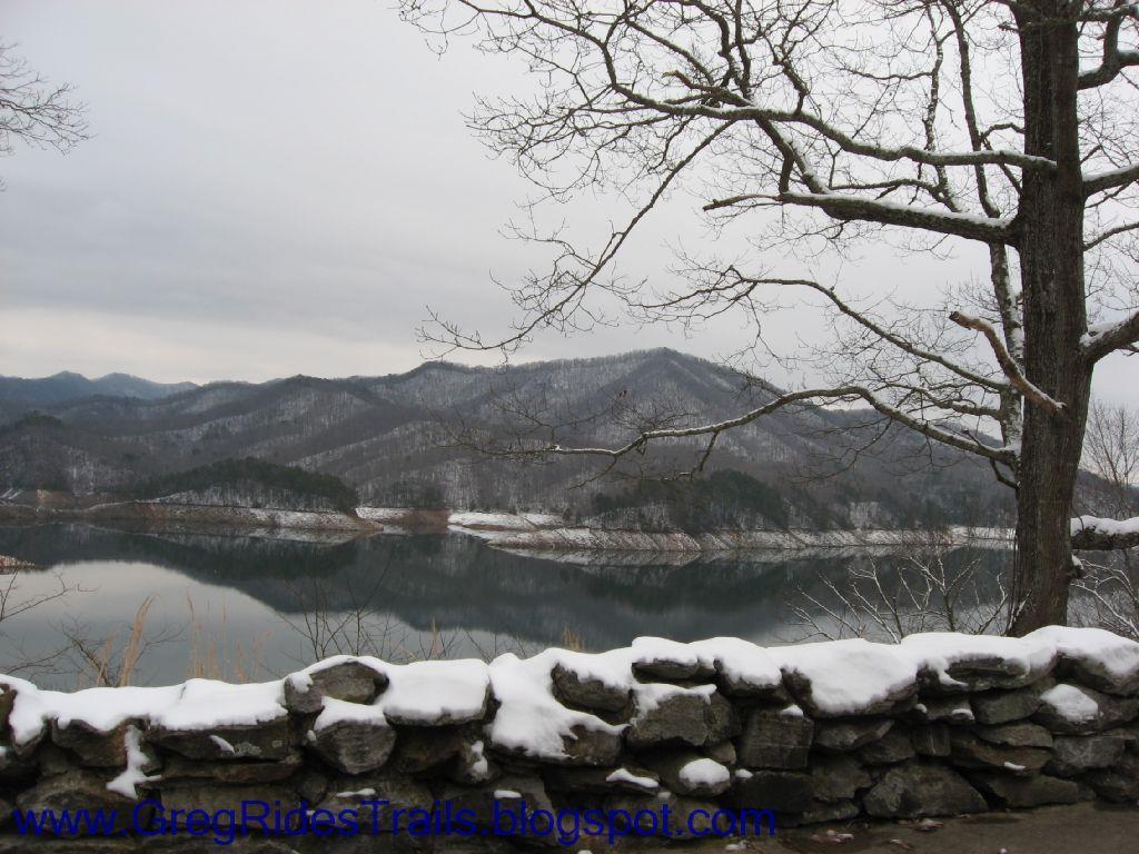 A serene winter landscape featuring a calm lake reflecting the surrounding snowy mountains under a cloudy sky. Bare trees are visible in the foreground, alongside a stone wall. The scene evokes a tranquil, peaceful atmosphere. Fontana Village mountain bike trail.