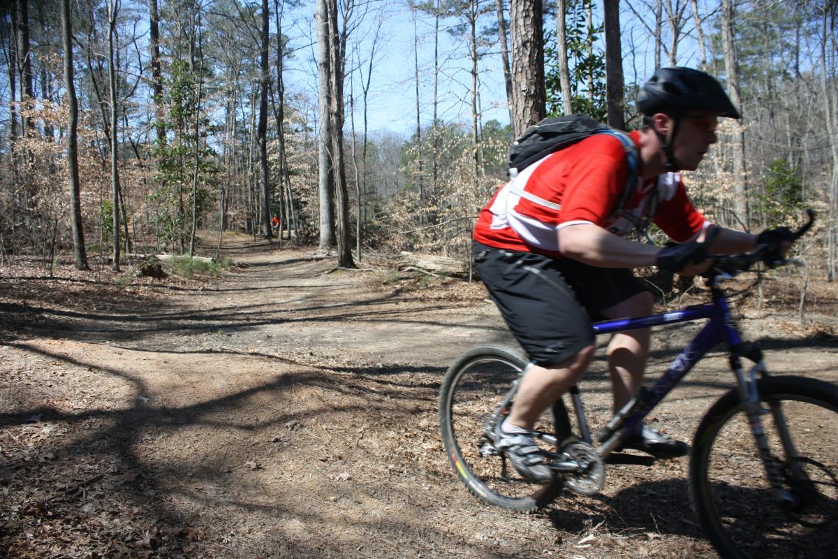 A person riding a mountain bike on a dirt trail through a wooded area, with trees and fallen leaves visible in the background. The cyclist is wearing a red jersey and a black helmet, and is moving at a moderate speed. In the distance, another person can be seen on the trail. Sope Creek mountain bike trail.