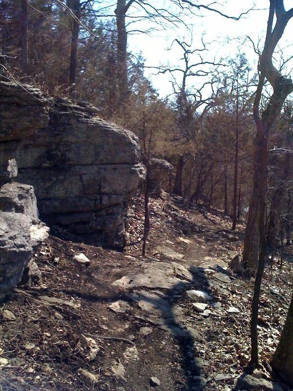 A narrow, rocky trail winds through a forested area during daylight. Large stone outcrops are visible on the left side, with scattered rocks along the path. The trees are mostly bare, indicating it may be early spring or late autumn. The sunlight casts shadows on the trail, creating a peaceful, natural setting. Swope Park Trail mountain bike trail.