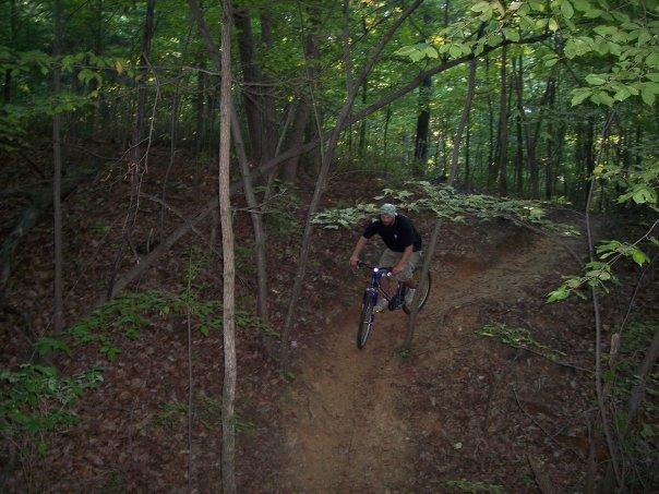 A person riding a mountain bike down a dirt trail in a wooded area, surrounded by green trees and foliage. The bike is in mid-motion, capturing the action of biking downhill on a natural terrain. Liberty Mountain mountain bike trail.