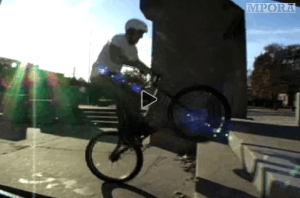 A cyclist performing a trick on a bicycle, lifting the front wheel off the ground while navigating outdoor concrete steps. The sun is shining brightly in the background, creating lens flare effects in the image.