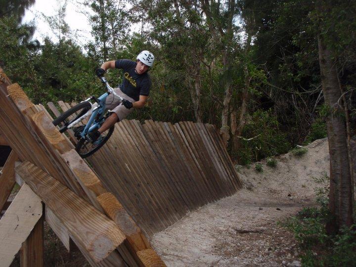 A mountain biker in a helmet and a t-shirt performs a trick on a wooden ramp in a forested area. The bike is tilted as the rider navigates a corner on the ramp, surrounded by trees and a sandy path. Markham Park mountain bike trail.